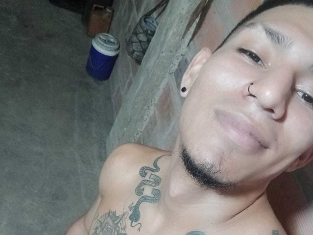 PeterJonies with a nose ring, black ear studs, short dark hair, and a goatee, showing tattoos on his chest and neck, shirtless, with a can in the background.