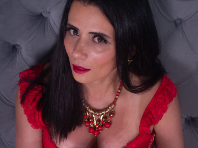 DaneshkaNaughty69 with dark hair, red lipstick, and red lace top, showing her big tits, wearing a red and gold bead necklace, looking up with a slightly seductive smile.