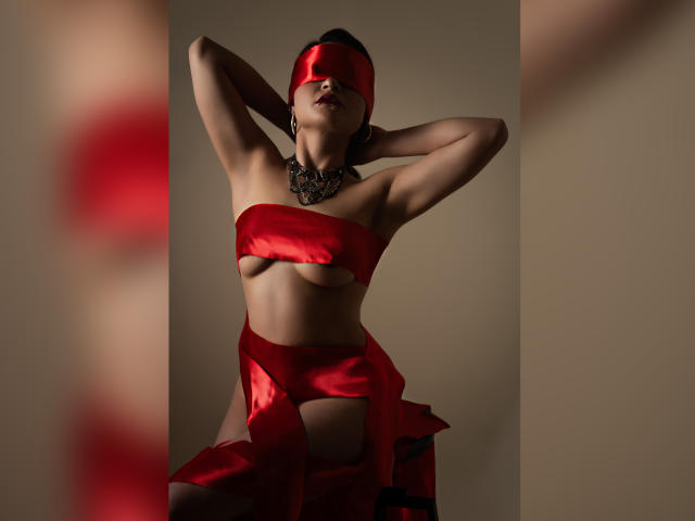 CassieJones with blindfold, red satin wrapping her tits and ass, hands behind head, necklace, shadowy light, seductive pose.