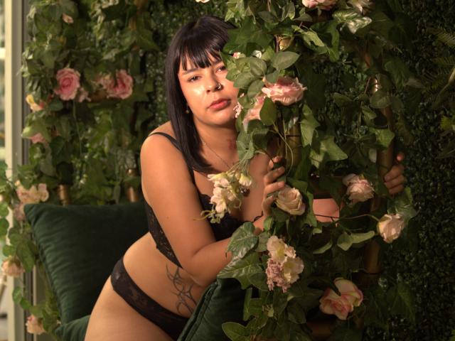 KylieDark with black lace bra and panties, holding roses, has tan skin, dark hair, and a spider web tattoo on her side, leaning against a green, floral-covered wall.