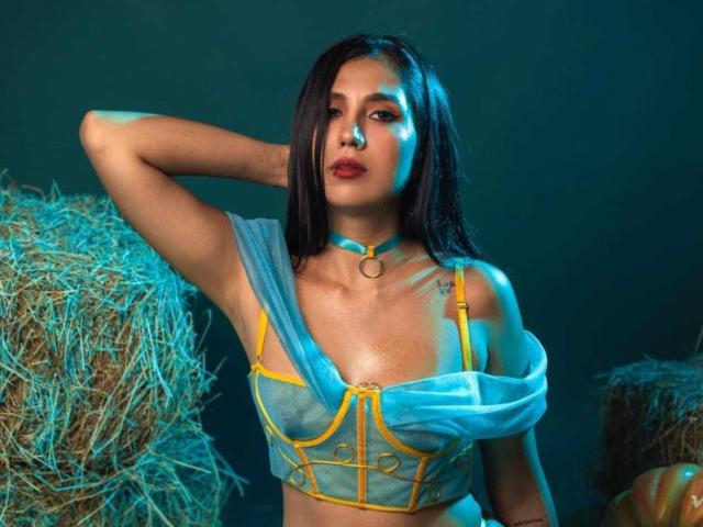 MissNatashaa with wet dark hair, wearing a blue choker, translucent yellow and blue top, exposing her small tits, arms up, next to hay bales under blue lighting.