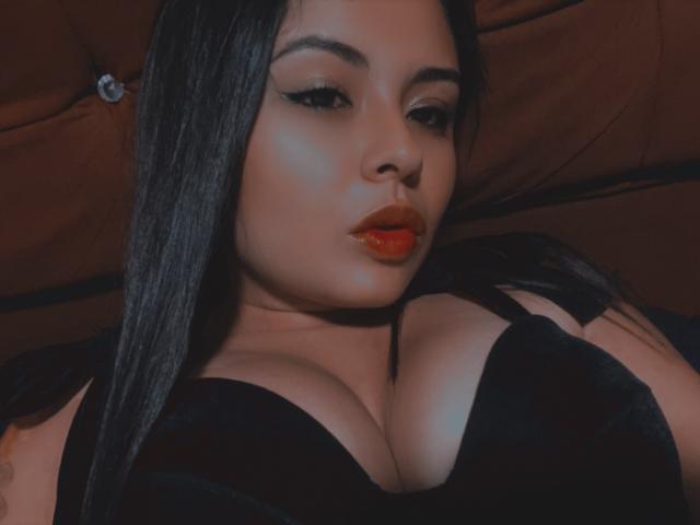 ViolethhLee has big tits in a black bra, with long black hair, red lipstick, and winged eyeliner, lying back with a brown headrest in the background.