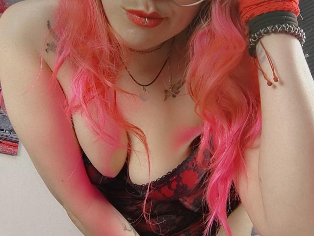 NatashaLau with pink hair, red lipstick, glasses, black lace top, showing her tits, wearing red gloves, a necklace, and a wrist bracelet, leaning forward.