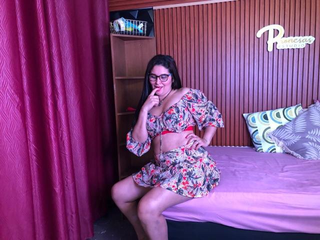 KarinaParker with glasses, floral crop top, and matching skirt, sitting on pink bed, finger in mouth, red belt, midriff, curvy, butt, dark hair, wood panel wall, pink curtain.