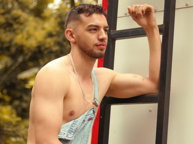 MichaelMagno, shirtless with denim overalls, leans on a ladder. He's got short curly hair, a beard, nose ring, and a chain necklace. His arm tattoo is visible.