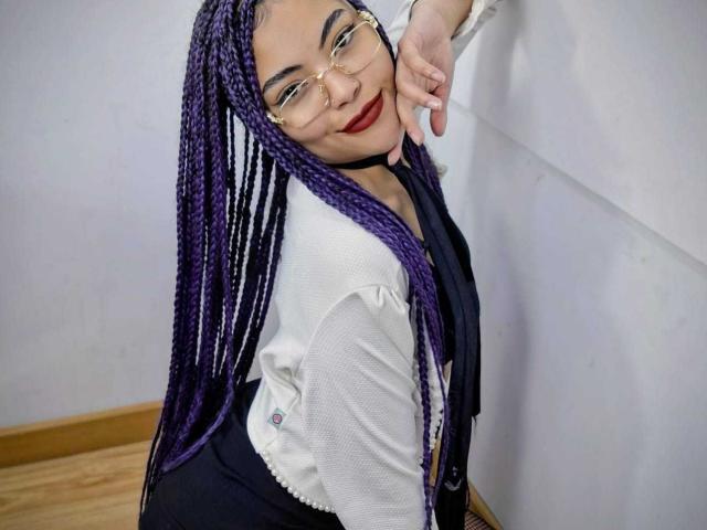 SammyHills69 with purple braids, glasses, white shirt, black choker, black top, and red lipstick, leaning against wall, smiling, showing her ass, and resting her hand on her cheek.