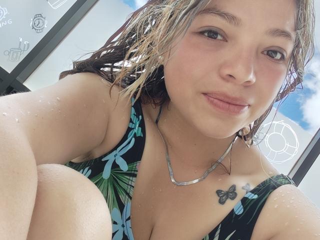 ScarlettAmelia with wet, brown, wavy hair, wearing a black, blue, and green floral swimsuit, showing her small butterfly tattoo on the right shoulder, and a silver necklace.