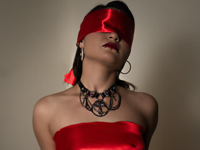 CassieJones with blindfold, red satin top, black chain necklace, red lipstick, hoop earrings, and red feather in her hair, head tilted back, eyes covered.