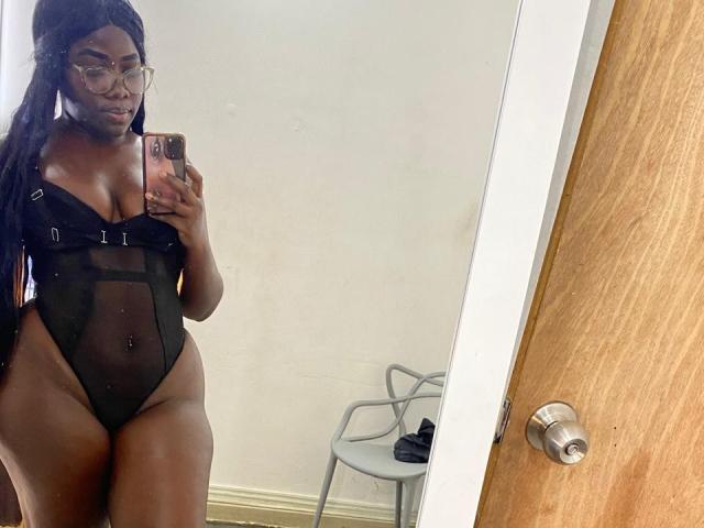 SweeterChocolate with dark skin, long braids, glasses, wearing a black mesh one-piece showing her tits and pussy, taking a mirror selfie in a room.