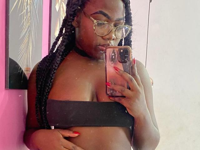 SweeterChocolate with dark skin, braided hair, gold glasses, black choker, and red nails, taking a mirror selfie, showing her boobs in a black strapless top.