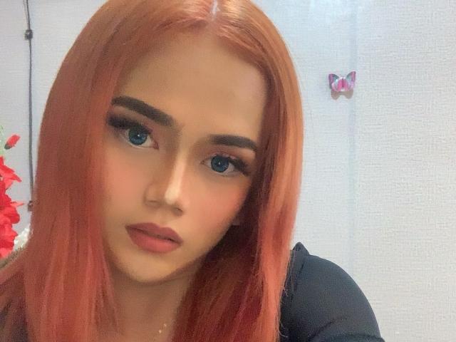 LustHaven has long red hair, blue eyes, full eyebrows, and pink lips. She's wearing a black top, looking at the camera with a neutral expression.