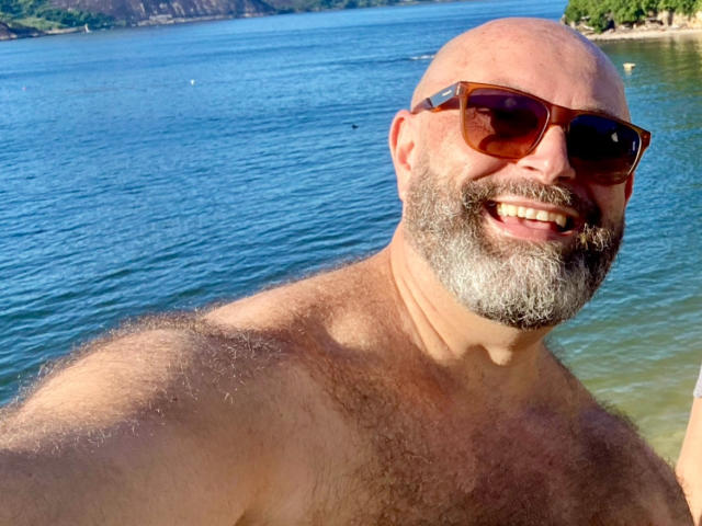 DmitriBr with a bald head, gray beard, and brown sunglasses, shirtless, smiling at the camera, showing a hairy chest, standing by a blue, calm water body.