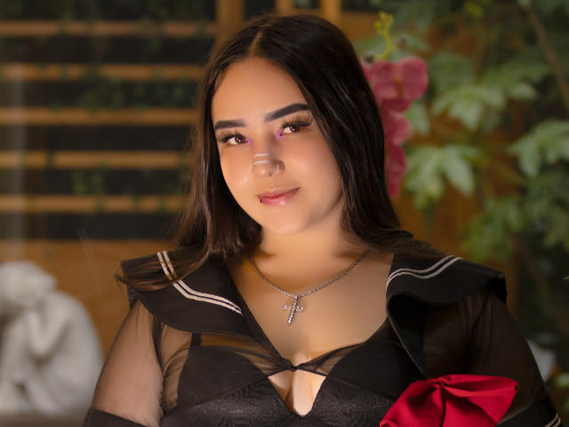 Morgan Hill, com cabelo castanho escuro, vestindo um uniforme escolar preto transparente, mostrando seus seios através de um recorte no decote, um laço vermelho e um colar com uma cruz, sorrindo suavemente..