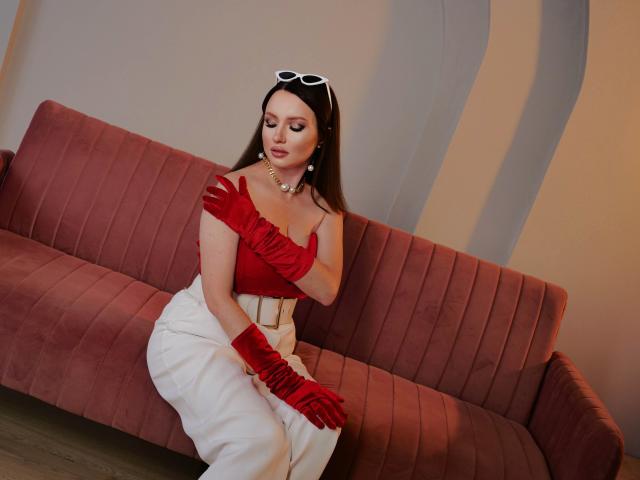 EndlessLovee sitting on a pink couch, wearing a red off-shoulder top, white pants, red gloves, and a pearl necklace, with white sunglasses on her head.