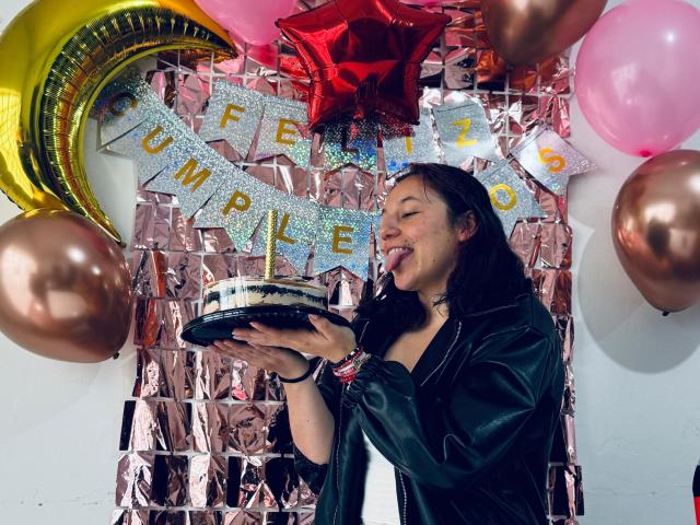 MilaWilde, with black hair and green shirt, licking a cake, surrounded by balloons and metallic decorations, tongue out, holding cake with candle.