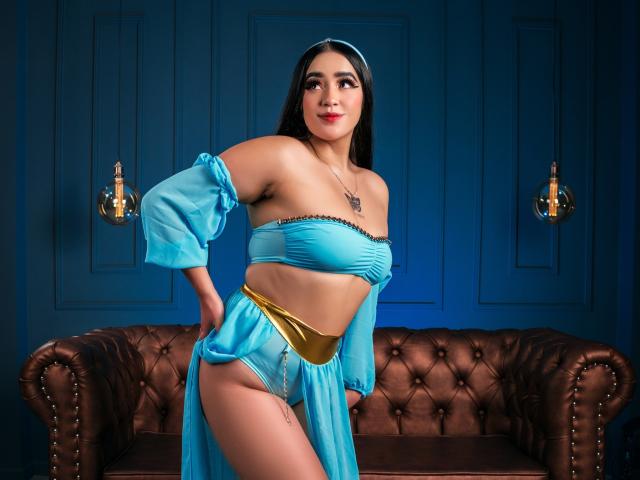 JennaStone with tan skin, black hair, blue strapless top, gold belt, sheer blue skirt, exposing her tits and pussy, standing in front of brown leather couch, smiling.