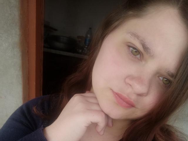 NettieElsie has light skin, green eyes, and reddish-brown hair. She's looking at the camera with a slight pout, her hand touching her chin. She's wearing a dark top.