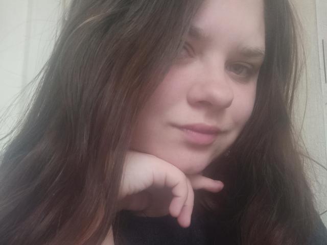 NettieElsie with wavy brown hair, pale skin, and a subtle smile, rests her chin on her hand, showing her nose and pink lips. Her eyes are slightly downcast.