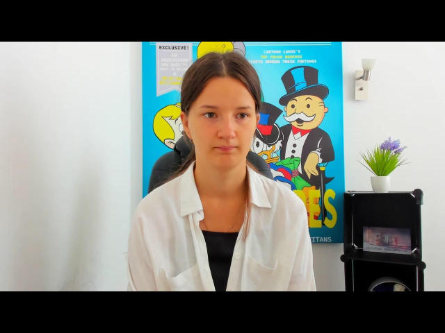 Didiene has a serious face, brown hair in a ponytail, wearing a white lab coat over a black shirt. A colorful poster with cartoon characters is behind her.