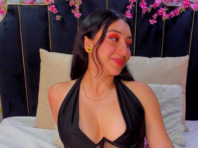 ArianaZahar with long black hair, red eyeshadow, yellow earrings, black halter top showing her big tits, smiling at the camera, sitting on a bed with beige pillows.