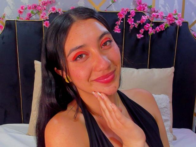 ArianaZahar has long black hair, red eye makeup, and a black halter top. She's smiling, touching her chin, with pink flowers on a black headboard. Her tits are covered.