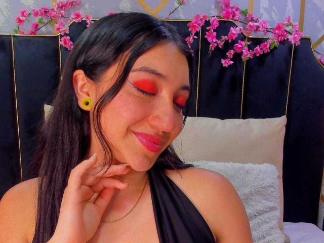ArianaZahar with black hair, red eye makeup, yellow ear gauges, black halter top, pink lipstick, smiling, hand near chin, pink cherry blossoms above black headboard.