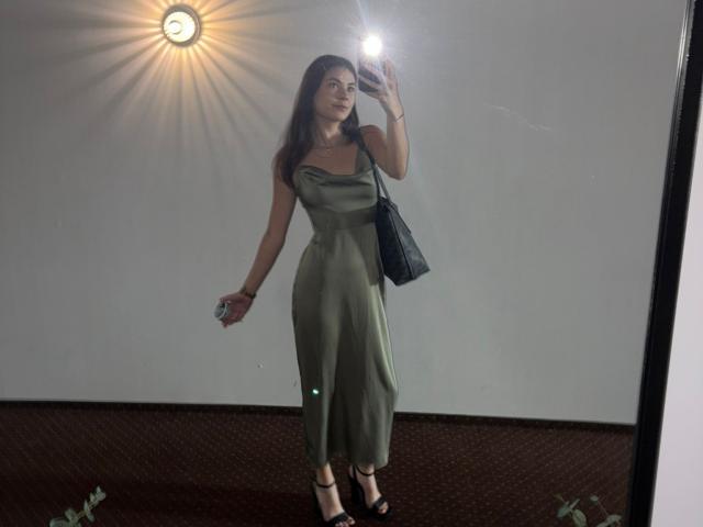 NancyGlow heeft borsten en draagt een groene, mouwloze, aansluitende jurk, zwarte hakken, ze houdt een telefoon vast, een spiegelzelfportret, een zwarte tas, ze staat, er is lichtreflectie, ze houdt een drankje in haar linkerhand..