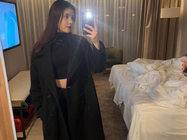 NancyGlow, met lang rood haar, draagt een zwarte coltrui en jas, houdt een telefoon vast en maakt een spiegel-selfie, waarbij haar slanke, mooie gezicht en kleine borsten te zien zijn..