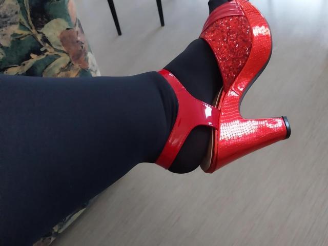 LiliChattePoilue's foot in a shiny red high heel with a textured glittery section, black tights, on a light wooden floor.