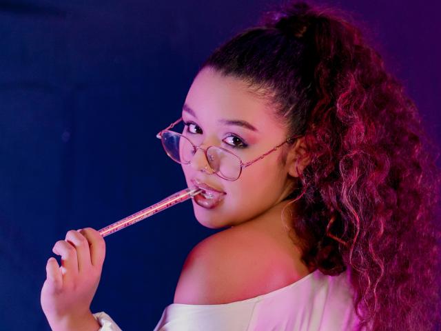 SofiaYue with curly pinkish-red hair in a ponytail, holding a pink glittery stick in her mouth, wearing off-shoulder white top, and pink glasses, looking back.
