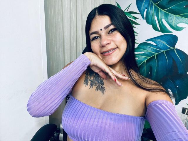 KeraniMarala with dark hair, purple off-shoulder top, small nose ring, tattoo on chest, smiling, hand on chin, purple nails, green leafy plant in background.