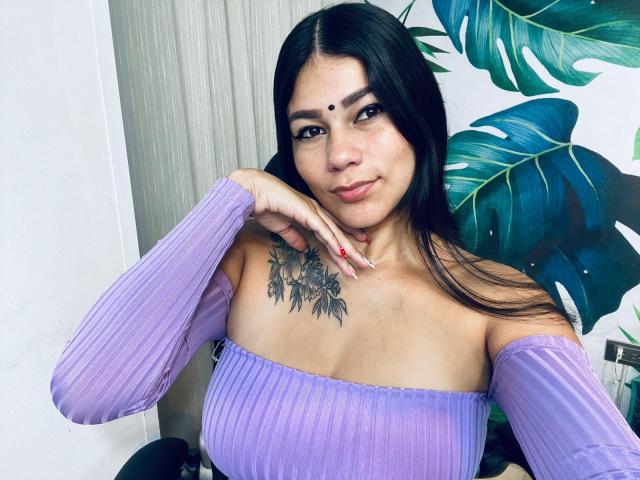 KeraniMarala met lang zwart haar, een paars top met een open schouder, een tatoeage op haar borst, rode nagellak en een stip op haar voorhoofd, lachend naar de camera..