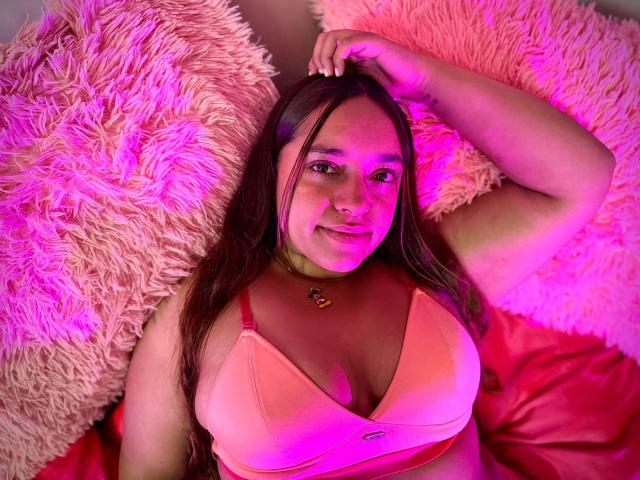SophiaLex with pink-lit boobs, laying on fluffy pink pillows, wearing a peach bra, hand in brown hair, smiling, pink light, brown eyes, cute, sexy, chest up.