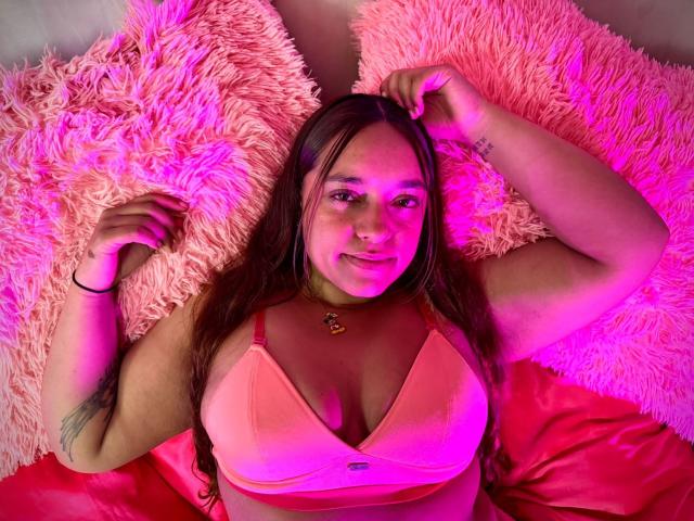 SophiaLex with pink bra, medium tits, brown hair, lying on pink fluffy pillows, pink and purple lighting, smiling, right arm raised, black bracelet, tattoo on left arm.