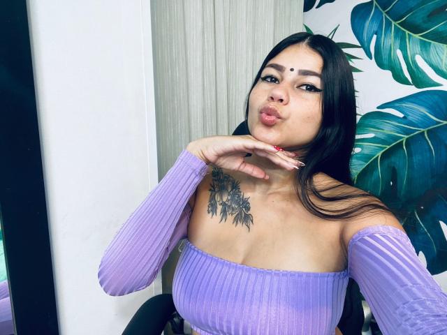 KeraniMarala, con labbra piene, capelli scuri, una maglietta viola con una spalla scoperta, un tatuaggio floreale sul petto, sopracciglia nere e una collana rossa, che si tocca il mento, mostrando il seno..