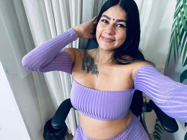 KeraniMarala, com cabelos longos e pretos, blusa roxa com detalhes em relevo e sem alças, e saia combinando, mostrando os seios e uma tatuagem de flor no peito, sorrindo, com a mão no cabelo..