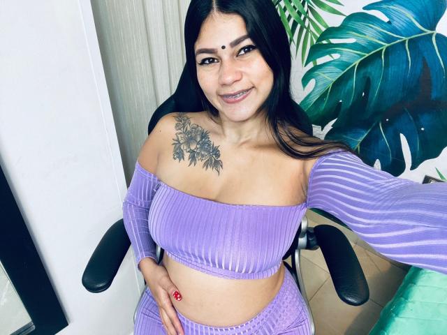 KeraniMarala with black hair, purple ribbed top, matching pants, tattoo on left shoulder, smiling, sitting, hand on hip, red nail, green leaf pattern in background.