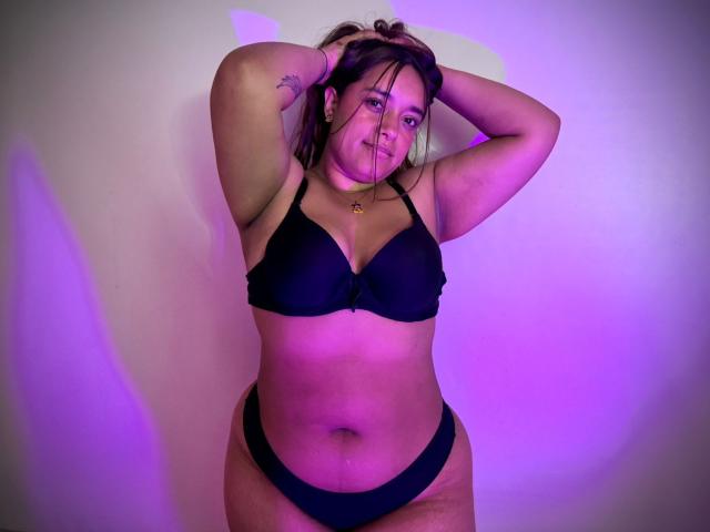 SophiaLex with arms raised, wearing black bra and panties, has a plus-size body, small tits, brown hair, tattoo on right arm, and a slight belly, under purple lighting.
