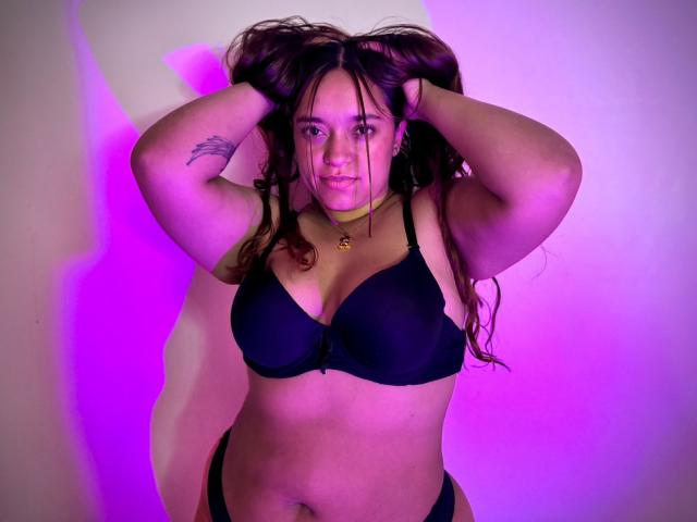 SophiaLex with curly brown hair, black bra, and black panties, hands in hair, tattoo on right arm, purple and pink lighting, slightly smiling, medium breasts, soft belly.