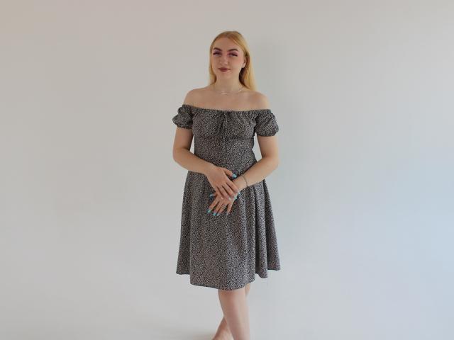 Beatrice: Een vrouw met blond haar, een grijze jurk met open schouders, kleine borsten, met haar handen samengevoegd, staand zonder schoenen, met een eenvoudige ketting en blauwe nagellak..