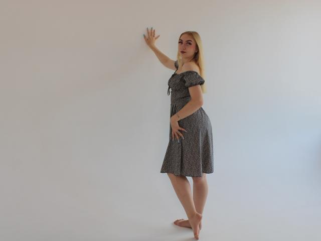 BeatriceFly stands barefoot, wearing a gray, off-shoulder dress, hand on wall, other hand on hip, looking sideways with long blonde hair, soft light, neutral background.