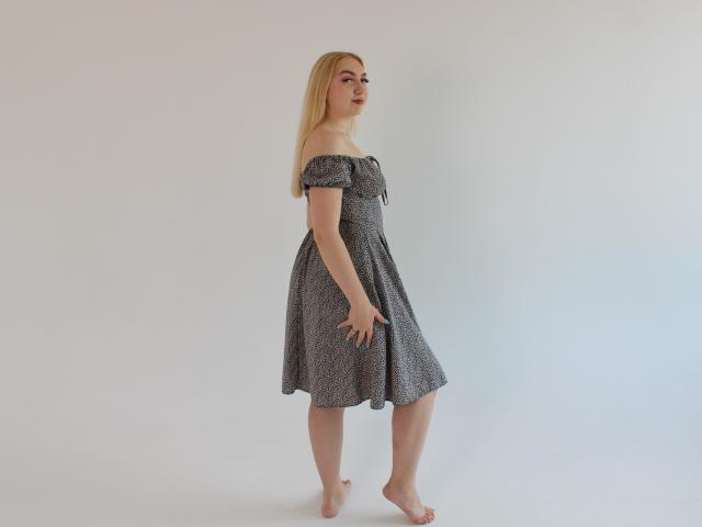 BeatriceFly se tient pieds nus, vêtue d'une robe grise à motifs, avec une épaule dénudée, et ses longs cheveux blonds. Elle regarde par-dessus son épaule, montrant ses petites fesses et ses petits seins..