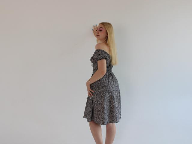 BeatriceFly, rubia, con sombra de ojos rosa, vestida con un vestido de lunares con un hombro descubierto, está de lado, tocándose las nalgas con una mano, mirando por encima del hombro con una expresión de puchero..