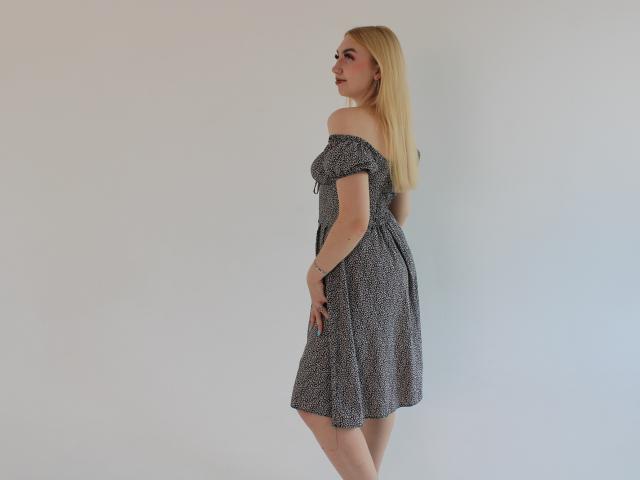 BeatriceFly with long blonde hair, off-shoulder grey dress, small tits, and a tattoo on her right wrist, standing side profile against a plain white wall.
