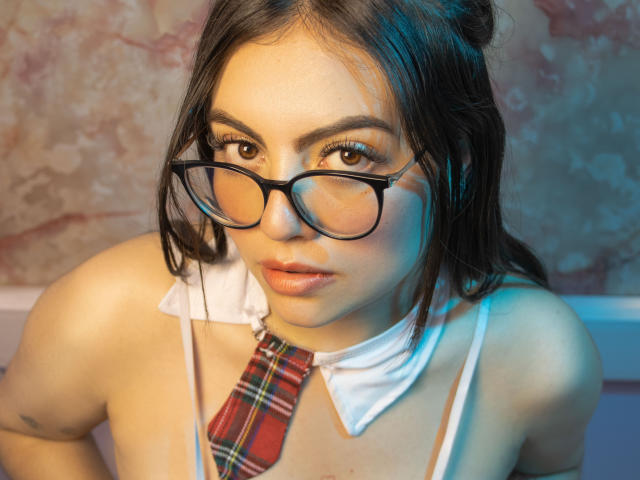 DorothyLane, met een bril, een geruit dasje en een witte kraag, kijkt omhoog, heeft volle lippen en donker haar, draagt een doorschijnende top, waardoor haar borsten zichtbaar zijn..