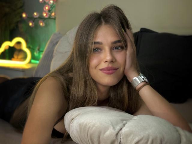 MelissaCrayzy, lying on a bed, with long brown hair, blue eyes, and pink lips, wearing a black top, resting her head on her hand, smiling gently, wearing a silver watch.