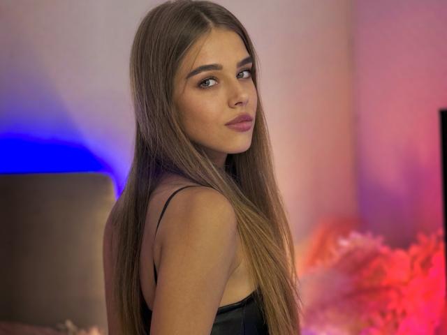 MelissaCrayzy with long brown hair, looking over her shoulder, wearing a black strap dress, showing her ass, and a subtle smile in a softly lit room.