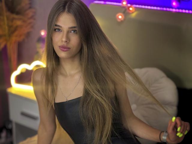 MelissaCrayzy with long, straight brown hair, wearing a black leather dress, standing with a slight smile, neon lights in the background, and a silver necklace. Boobs are small.