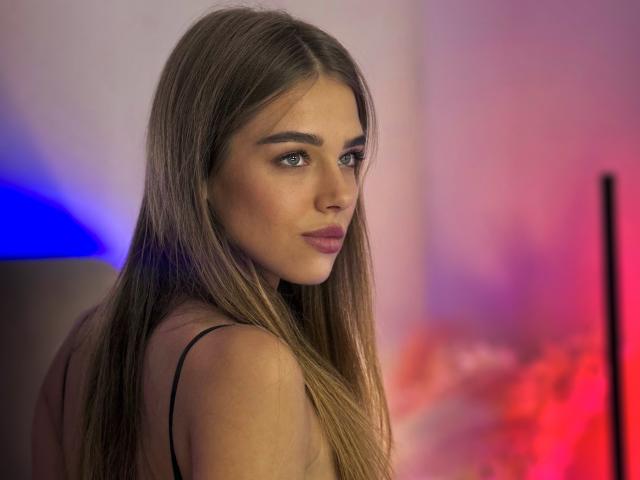 MelissaCrayzy has long brown hair, blue eyes, and pink lipstick, wearing a black spaghetti strap top, looking over her shoulder with a slight smile. Her tits are small.