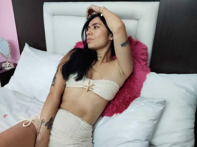 AlexaRosex with black hair, wearing a cream bandeau top and matching skirt, lying on a bed with a pink fuzzy pillow, right arm raised, showing tattoos.